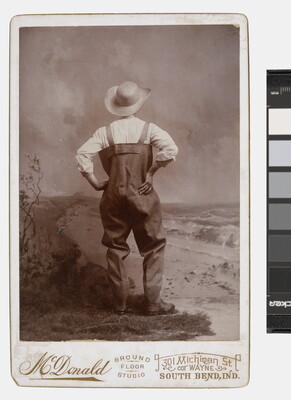 A sepia-toned studio photograph of a man, viewed from the back, wearing waders and a large straw hat, posed with hands on hips.