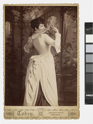 A sepia-toned studio photograph of a young White woman, viewed from the back, with a dramatically corseted dress and full bustle peeking playfully over her shoulder and hiding her face with a handkerchief.