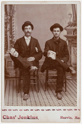 A black-and-white studio photograph of two seated White men, both wearing hats and three-piece suits, right legs crossed over left knees, holding cash in one hand.
