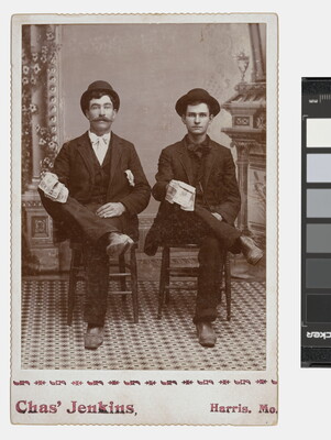 A black-and-white studio photograph of two White men seated next to each other in identical positions, wearing suits and hats, one leg crossed over the other.