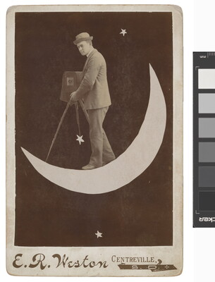A black-and-white photograph of a White man wearing a bowler hat and suit holding an old-fashioned camera on a tripod while standing on a crescent moon.