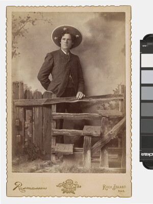 A sepia-toned photograph of a White man wearing a large sombrero-like hat standing behind a split-rail fence that is part of a studio set.