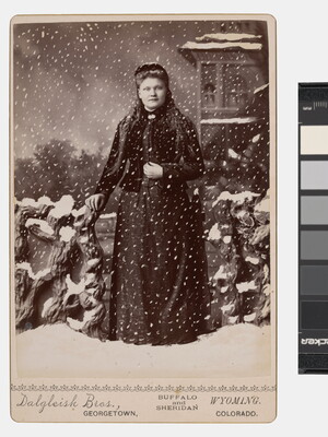 A black-and-white studio photograph of a White woman standing in a fake snowstorm against a backdrop of a winter scene.