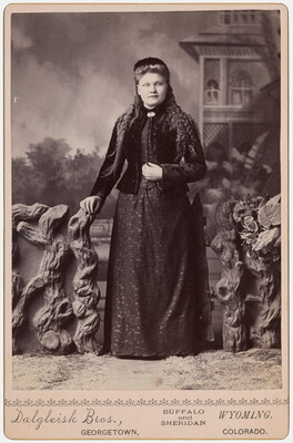 A black-and-white studio photograph of a White women dressed in black standing in front of a backdrop of an outdoor scene.