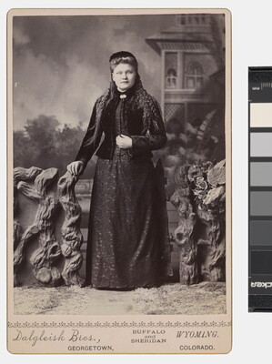 A black-and-white studio photograph of a White women dressed in black standing in front of a backdrop of an outdoor scene.