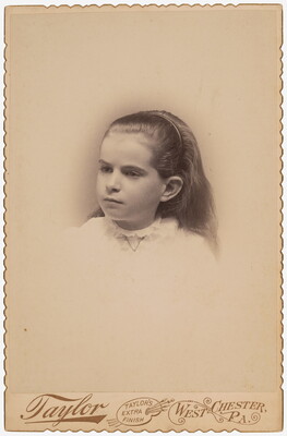 A black-and-white studio photograph of the head and shoulders of a young White girl with long hair held back in a headband.