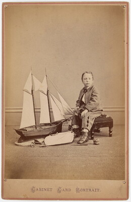 A sepia-toned studio photograph of a young White boy in a plaid suit seated on a bench next to a large toy sailboat.
