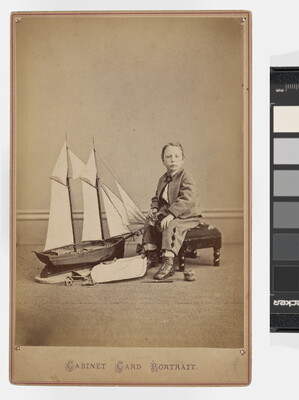 A sepia-toned studio photograph of a young White boy in a plaid suit seated on a bench next to a large toy sailboat.
