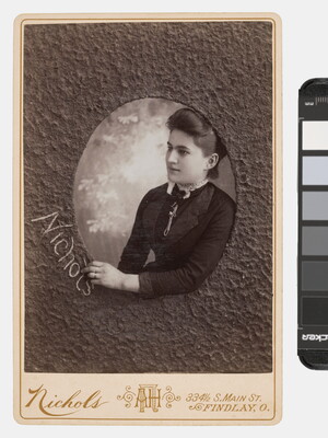 A sepia-toned studio photograph of a White woman who appears to be reaching through a textured oval window to write the word, "Nichols."