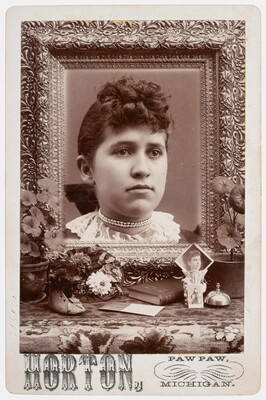 A black-and-white studio photograph of a young White woman, hair pulled back and a pearl choker around her neck; an ornate frame surrounds her portrait above a table with flowers and a book.