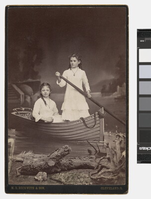 A sepia-toned studio photograph of two adolescent White girls, both wearing high-necked white dresses, posing in a rowboat in front of a painted lake scene.