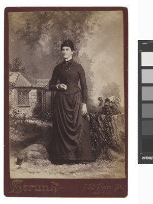 A sepia-toned studio photograph of a White woman with dark hair wearing a corseted, high-necked dress standing in front of a painted pastoral scene with a cottage.