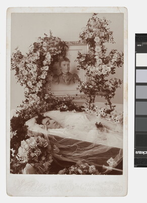A sepia-toned photograph of a deceased woman in a coffin covered with flowers and a framed photograph of her in life on top of the coffin.