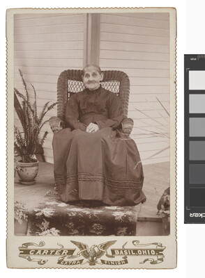 A sepia-toned studio photograph of an elderly White woman wearing a dark, high-necked dress sitting in a high back wicker chair on a porch with plants.