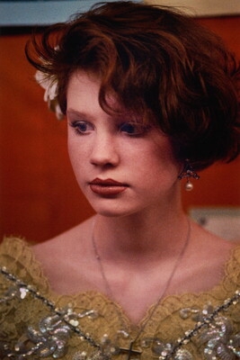 A color portrait photograph of a young White girl with short red hair, a flower tucked over one ear, and a decorative yellow top.