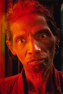 A color portrait photograph of a Black man cast in red light, hair combed back and stubble on his face, hoop earrings in one ear.