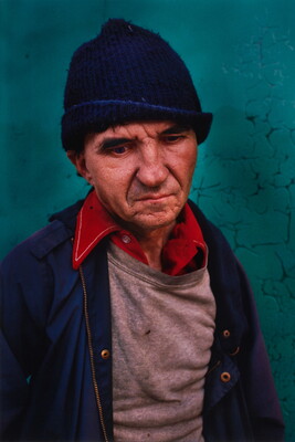 A color photograph of a middle-aged White man with deep wrinkles wearing a blue knit hat and a red shirt under a gray sweatshirt and blue jacket. 