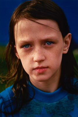 A color portrait photograph of a young White child with freckles, shoulder length brown hair, and blue eyes, wearing a blue knit sweater.