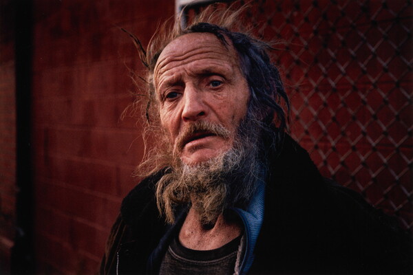 A color portrait photograph of an older White man with flyaway and matted hair, a graying beard and mustache, and a furrowed brow standing in front of a chain link fence.