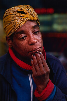 A color photograph of a middle-aged Black woman wearing a yellow headwrap and blue and red sweater holding her fingers to her chin.