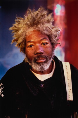 A color portrait photograph of an older Black man with matted gray hair and a beard wearing a black jacket.