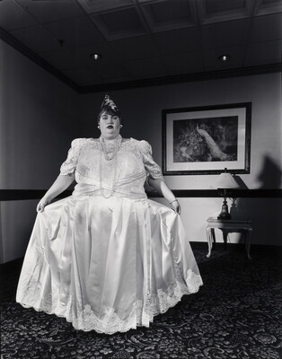 A black-and-white photograph of a White woman holding out the skirt of her long, formal white dress.