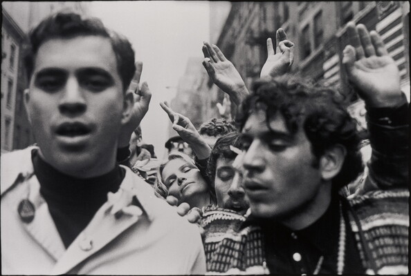A black-and-white photograph of two people amongst a crowd of protesters, some of whom are holding up their hands and making circles with their index fingers and thumbs.