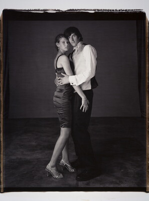 A black-and-white photograph of a young White couple in formal attire embracing one another and looking at the camera.