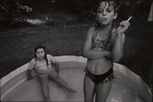 A black-and-white photograph of a young White girl standing in a kiddie pool smoking a cigarette as another young White girl sits in the pool behind her.