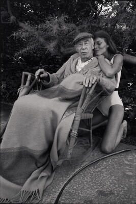 A black-and-white photograph of an older man seated in a rocking chair with a blanket on his lap as a younger woman kneels behind him to hug his shoulders.