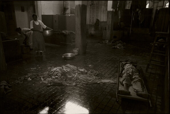 A black-and-white photograph of the inside of a room showing a pile of rags on a wet tile floor, people on one side of the room holding tubs, and a person lying on a cot on the other side.