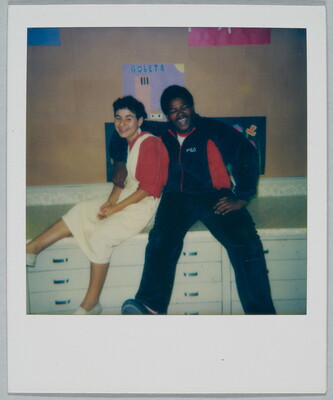 A color Polaroid photograph of a young, smiling White woman wearing a white dress over a red shirt seated on a cabinet next to a smiling Black man wearing a track suit with his arm around her.