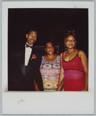 A color Polaroid photograph of a young Black man wearing a tuxedo with a red corsage, an older Black woman wearing a multi-colored shirt, and  a young Black woman wearing a formal red dress.