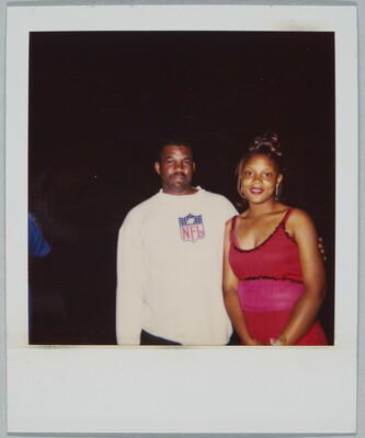 A color Polaroid photograph of a Black man wearing a white NFL sweatshirt with his arm around a young Black woman wearing a formal red dress.