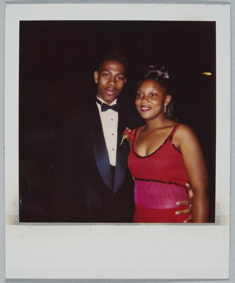 A color Polaroid photograph of a young Black man wearing a tuxedo with a red corsage with his arm around the waist of a young Black woman wearing a formal red dress.