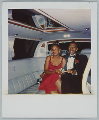 A color Polaroid photograph of a young Black woman wearing a formal red dress and a young Black man wearing a tuxedo with a red corsage holding hands inside a limousine.