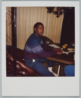 A color Polaroid photograph of a young Black man wearing a blue, white, and purple sweater seated at a table playing dominoes.