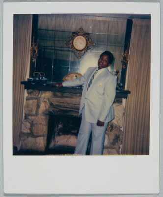 A color Polaroid photograph of a young Black man wearing a light gray suit and tie posing in front of a fireplace with his hand on the mantel.