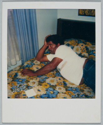 A color Polaroid photograph of a Black man wearing a white t-shirt and jeans lying across a bed with a blue and yellow floral bedspread.