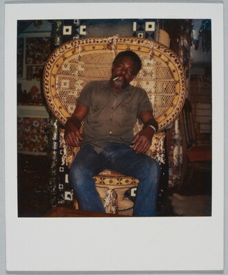 A color Polaroid photograph of a middle-aged Black man smoking a cigarette, wearing a button-down shirt and jeans, sitting in a wicker chair with wide back.