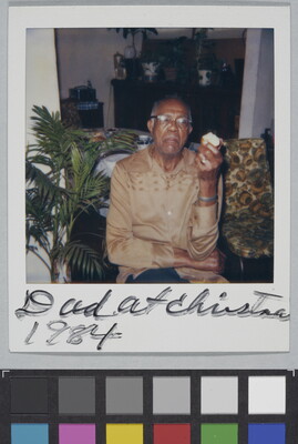 A color Polaroid photograph of a seated, middle-aged Black man in glasses and a light brown shirt holding an apple in one hand; handwritten text at bottom reads: "Dad at Christma[s] 1984."