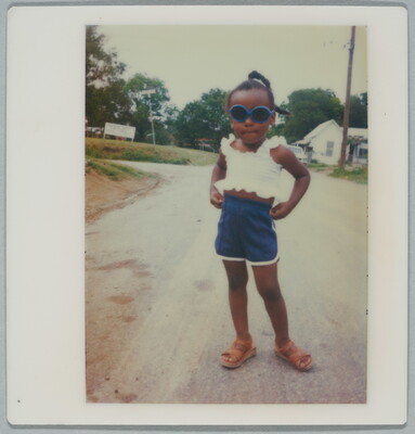 A color Polaroid photograph of a young Black girl wearing blue sunglasses, a white crop top, blue shorts, sandals and posing with hands on her hips.