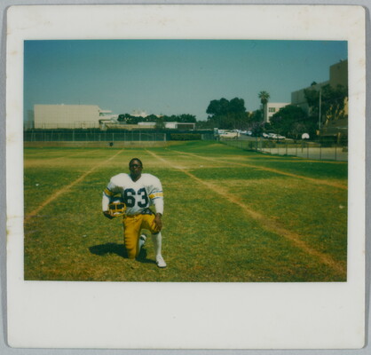 A color Polaroid photograph of a young Black man wearing a football uniform, holding his helmet under one arm, kneeling in a football field.
