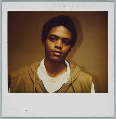 A color Polaroid photograph of a young Black man, head slightly tilted, wearing a hoodie over a white shirt.