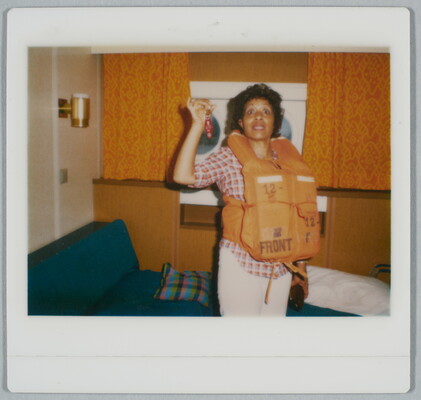 A color Polaroid photograph of a smiling Black woman wearing an orange life vest over a plaid shirt and white pants; in one hand she holds up keys dangling from a red keychain.