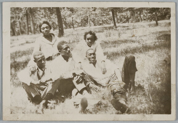 A black-and-white snapshot photograph of a group of Black people, three men and two women, seated in a grassy area with trees in the background.