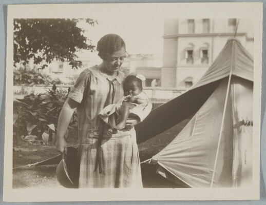 A black-and-white snapshot photograph of a Black woman wearing a plaid dress holding a baby in front of what looks like a tent.
