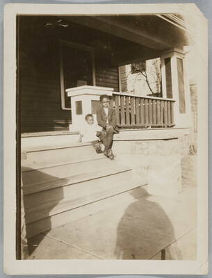 A black-and-white snapshot photograph of two Black children, one wearing a suit, sitting on the steps of a porch; the photographer's shadow on the sidewalk.