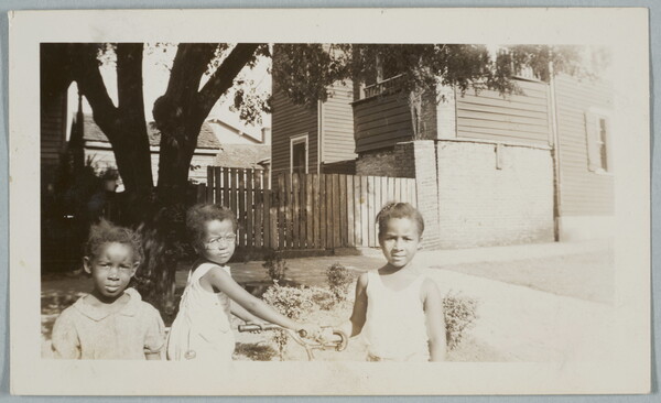 A black-and-white snapshot photograph of three Black children from the waist up playing in a yard; one holds the handles of a bicycle.