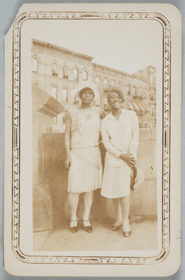 A black-and-white snapshot photograph of two Black women, both dressed in 1920s-style dresses and hats, standing in front of tall buildings.